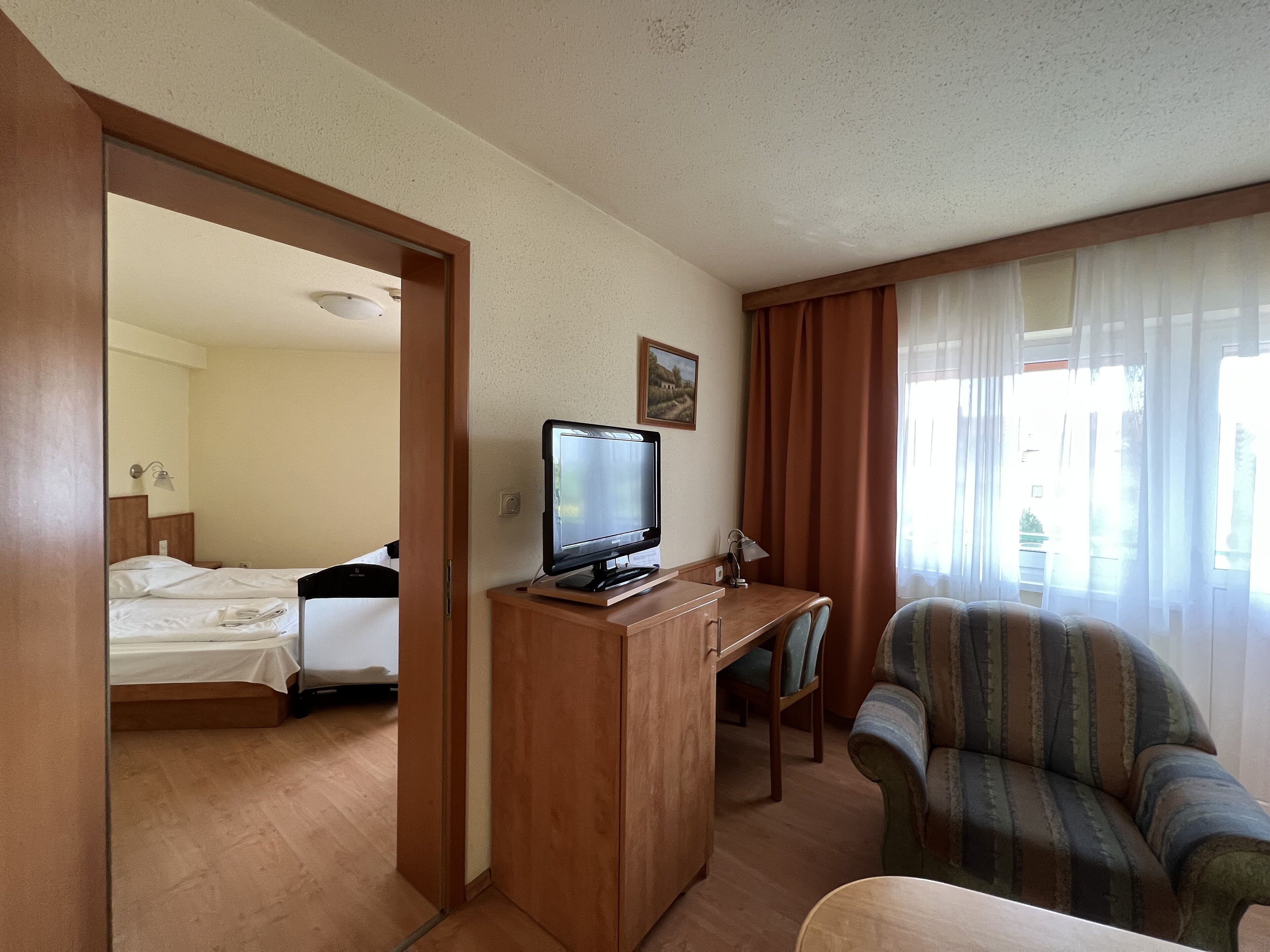 A hotel room with a bed, a TV on a wooden stand, a desk with a chair, and a window with curtains.