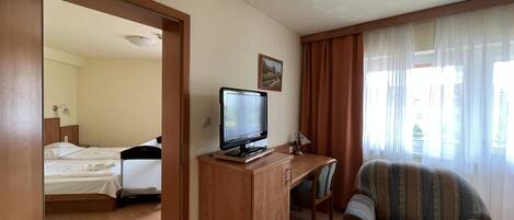 A hotel room with a bed, a TV on a wooden stand, a desk with a chair, and a window with curtains.