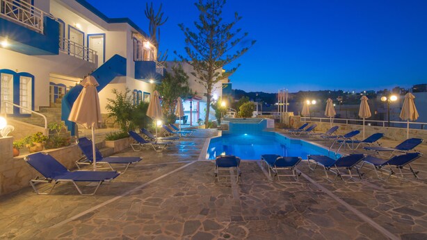 A pool area with lounge chairs and umbrellas, a stone floor, and a building with blue accents and a balcony. - Belvedere Village (Malevizi)