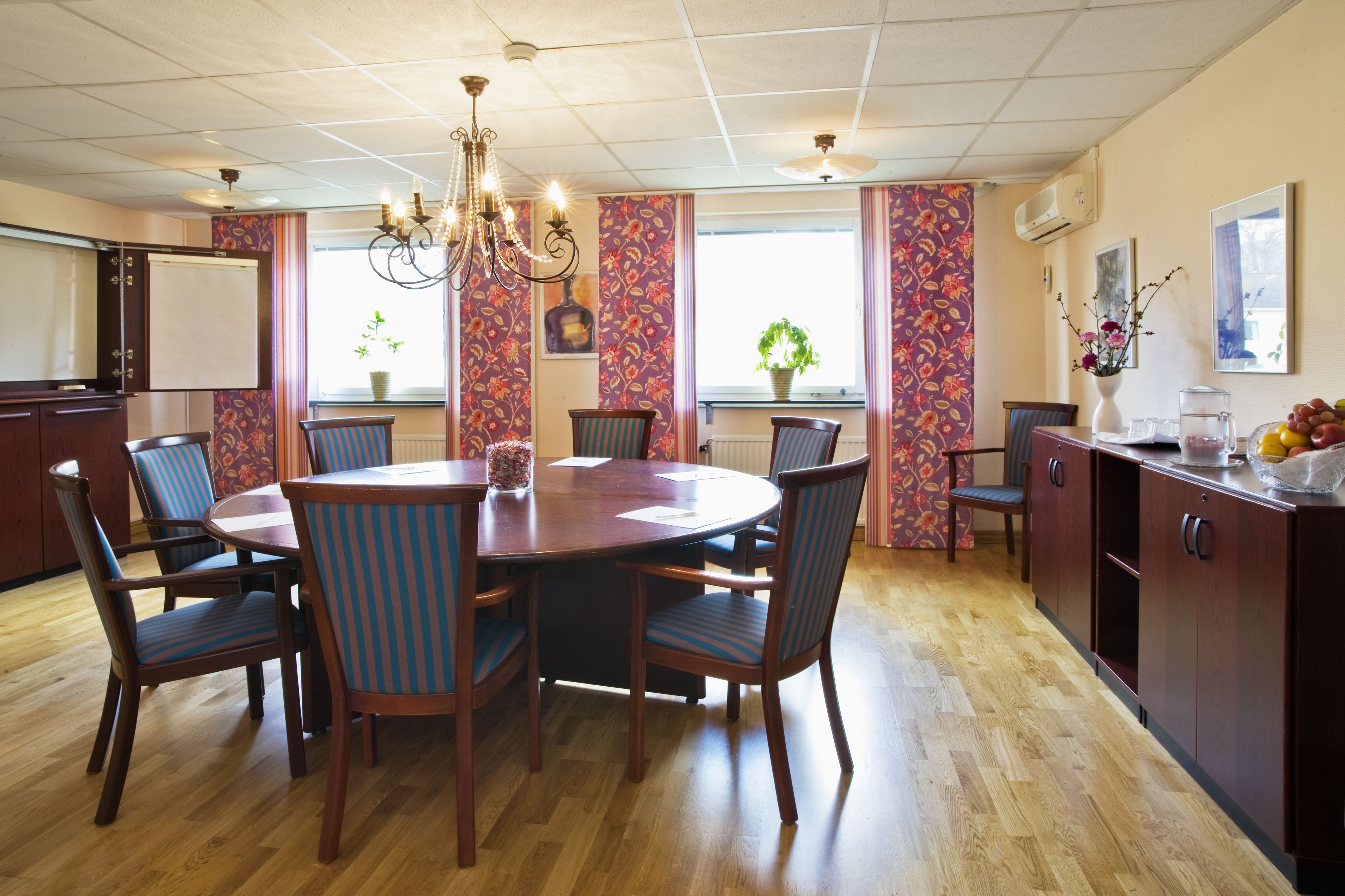 Un coin repas avec une table ronde en bois, six chaises, un lustre et une vue sur l’extérieur grâce à de grandes fenêtres ornées de rideaux à motifs floraux.