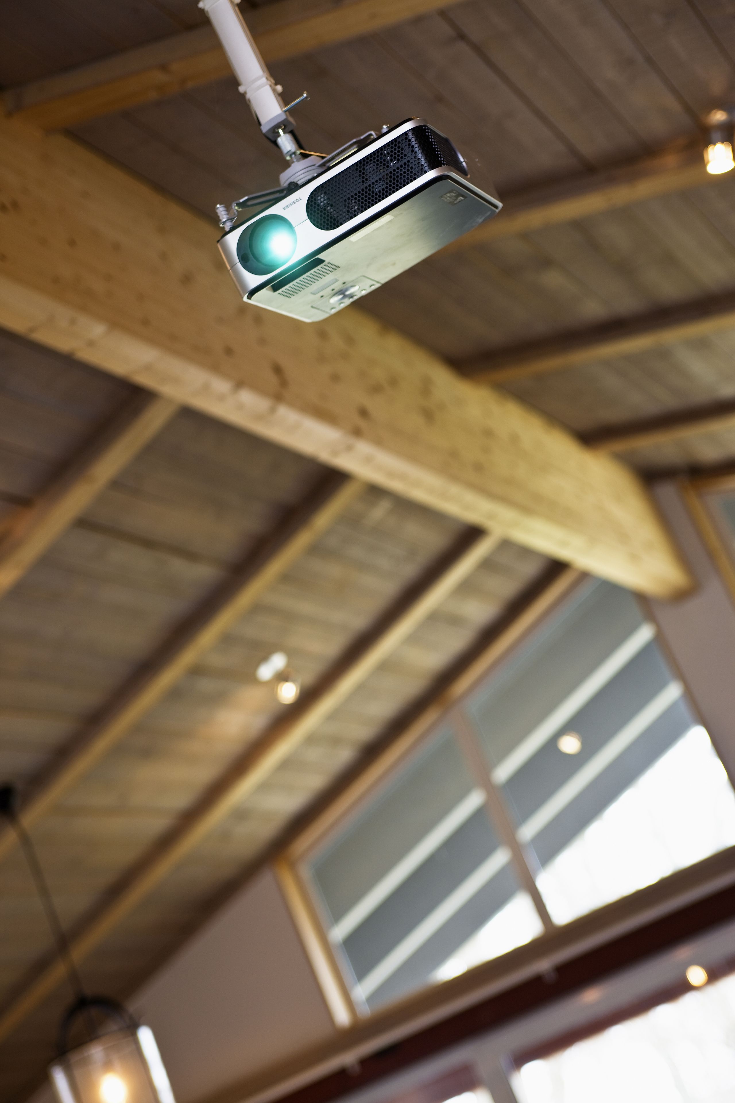 A ceiling-mounted projector in a room with wooden beams and large windows.