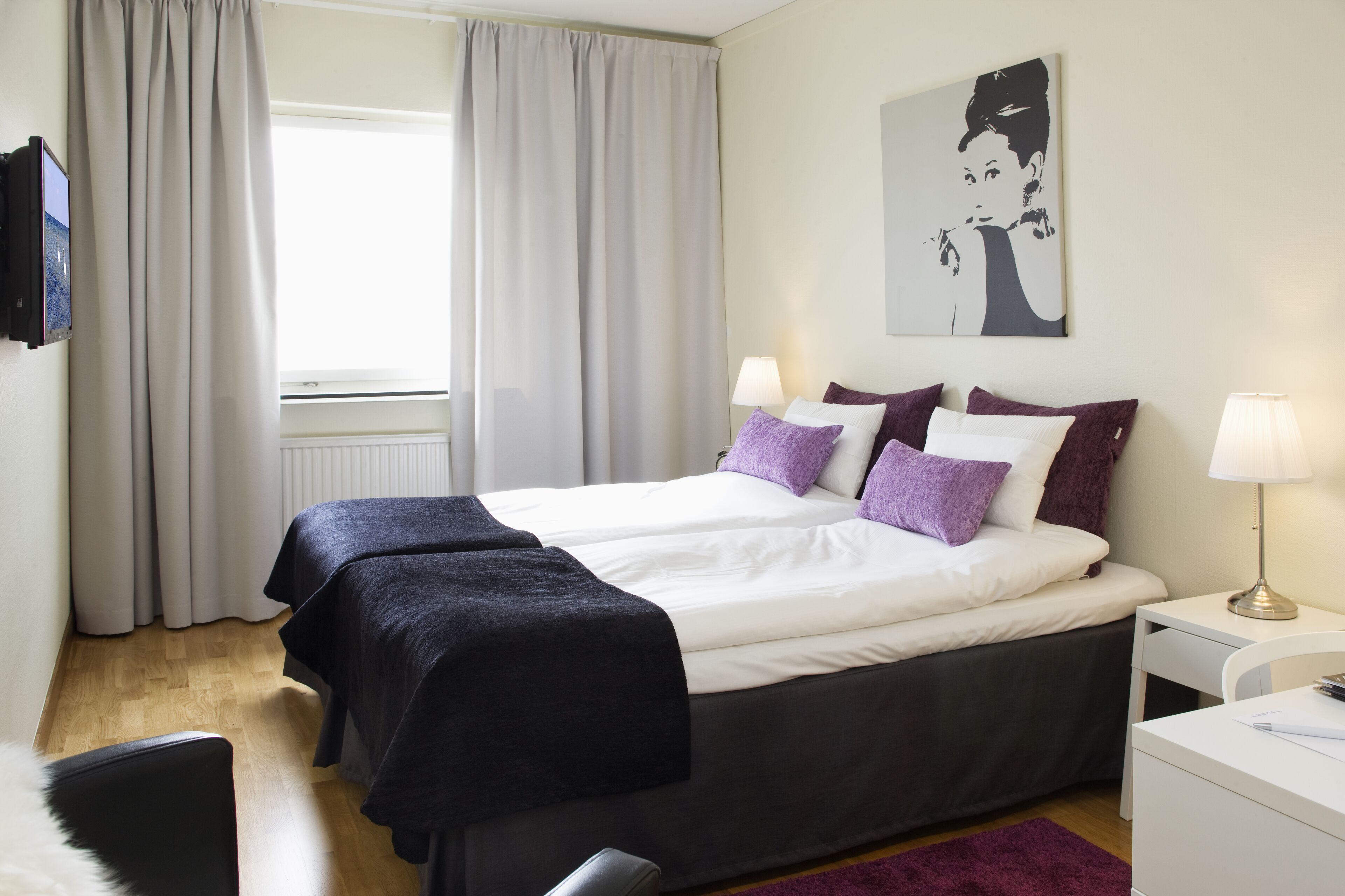 A hotel room with a bed, two bedside tables, a desk, a chair, a TV, and a window with curtains.