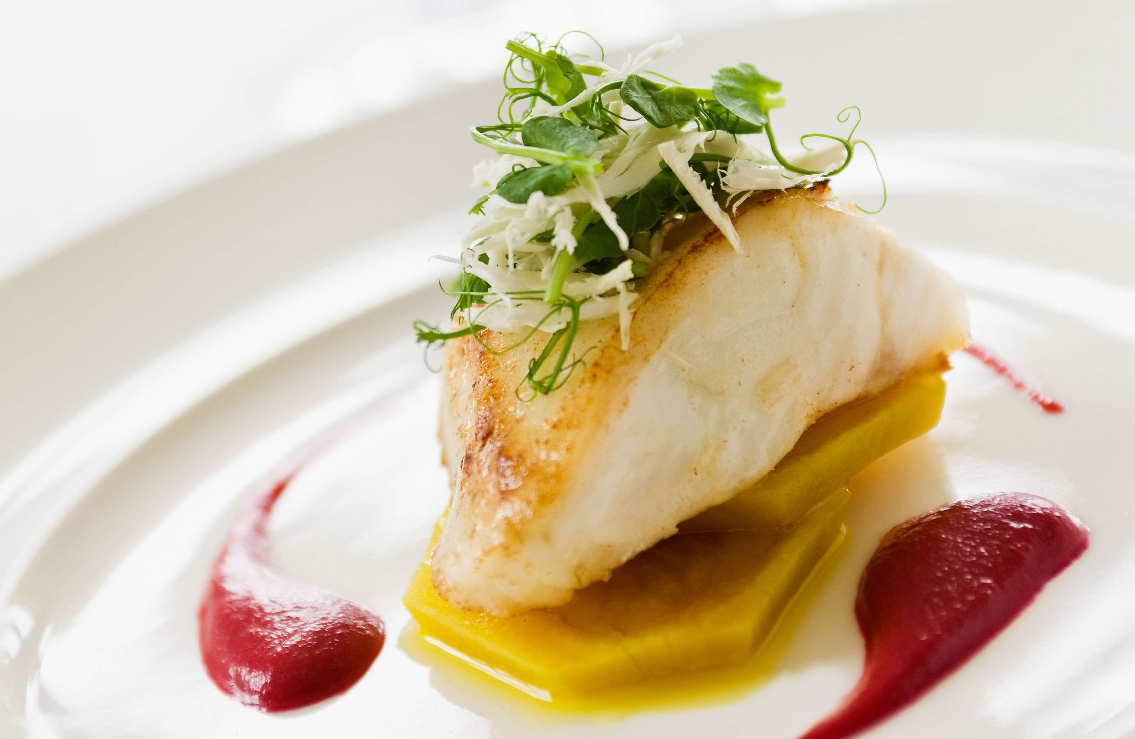 Un plato blanco con un trozo de pescado blanco cubierto de vegetales verdes y salsa amarilla.
