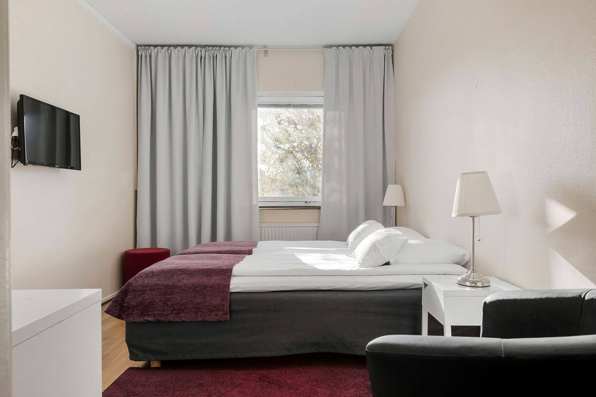 Een hotelkamer met een bed, een aan de muur bevestigde televisie, een bureau met lamp en een raam met gordijnen.