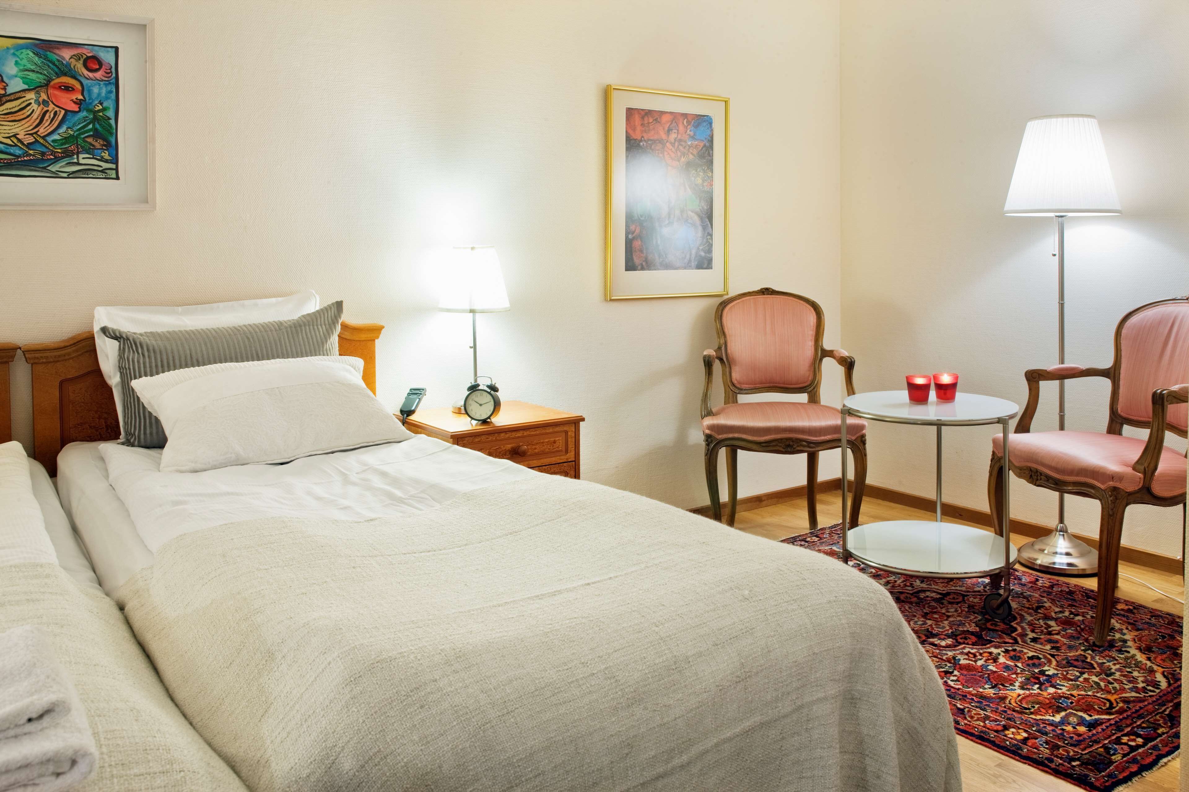A hotel room with a bed, two chairs, a small table, a lamp, and a clock on a nightstand.