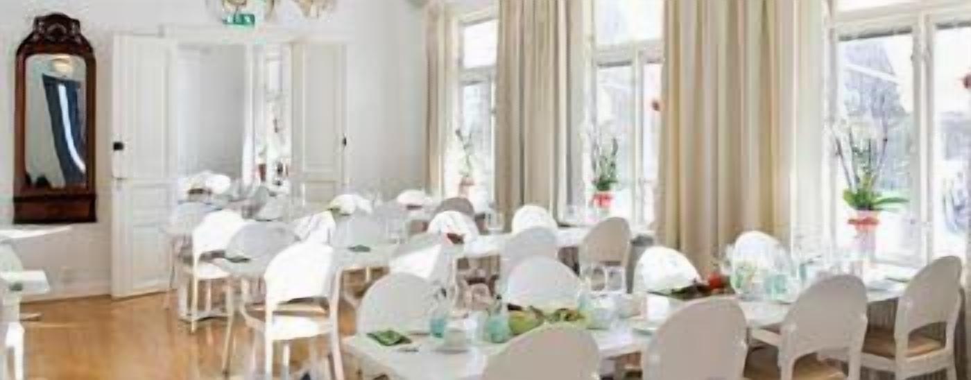 A dining area with multiple white tables and chairs, large windows with curtains, and a mirror on the wall.