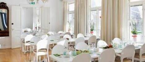 A dining area with multiple white tables and chairs, large windows with curtains, and a mirror on the wall.