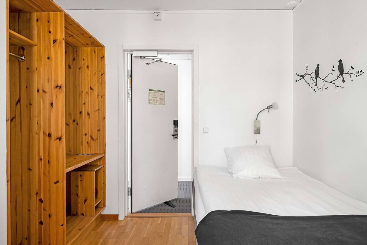 Een kamer met een houten garderobe, een bed met witte lakens, een wandlamp en een decoratie van vogels op een tak.
