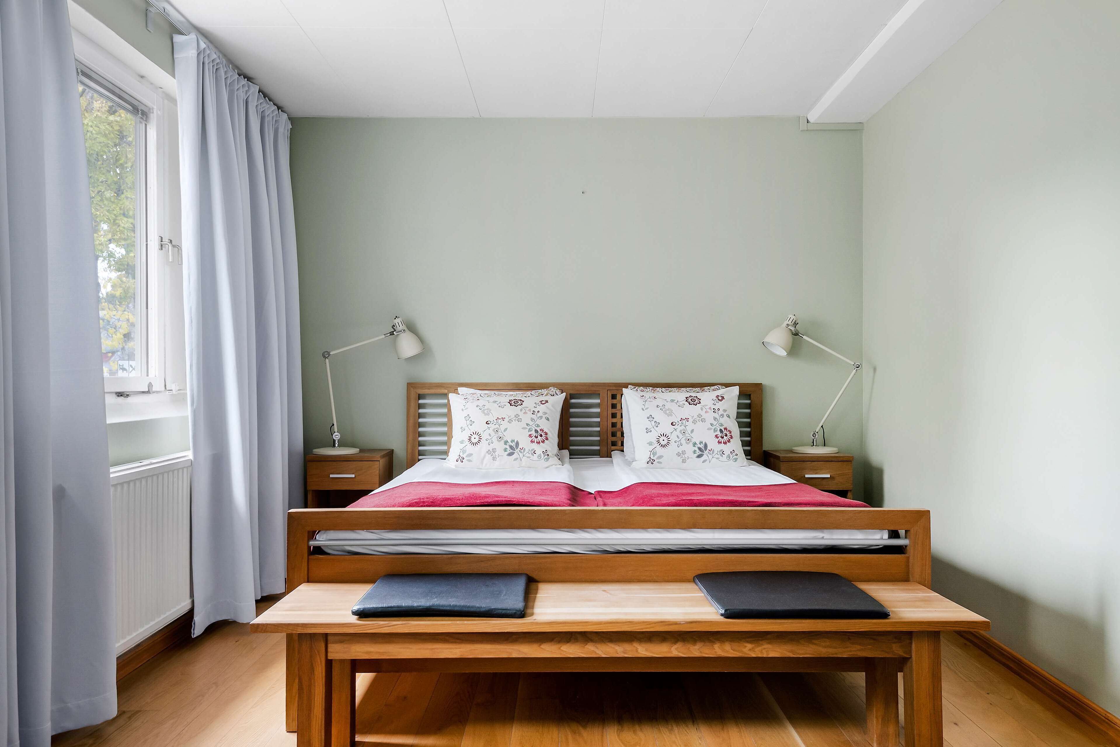A bedroom with a wooden bed, two bedside tables with lamps, a desk, and a window with curtains.