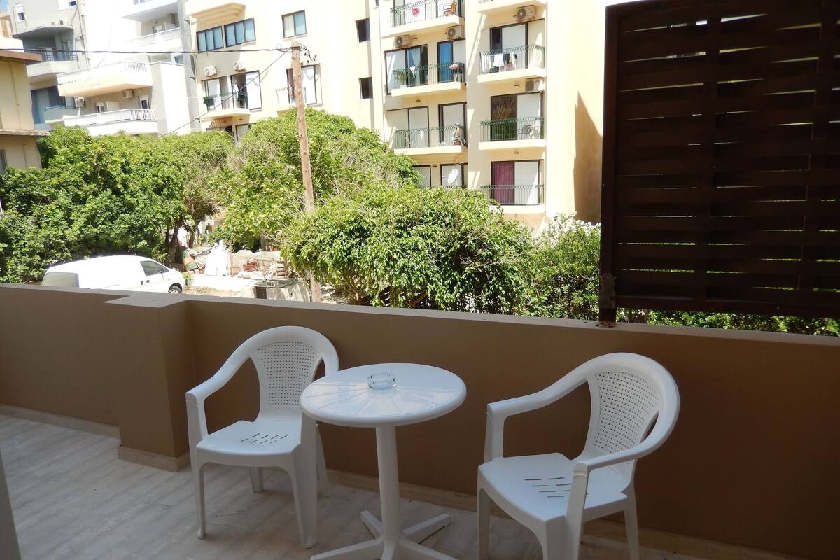 A balcony with two white plastic chairs and a small round table, overlooking a residential area with multi-story buildings and greenery.