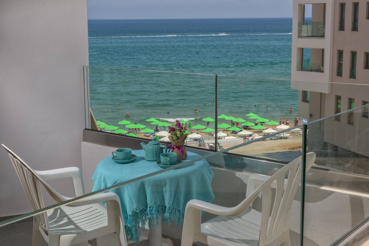 A balcony with a table set for two, overlooking a beach with umbrellas and people.
