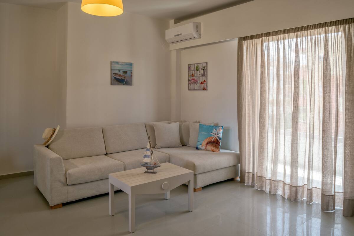 A modern living room with a beige sofa, a small white table, and a large window with sheer curtains.