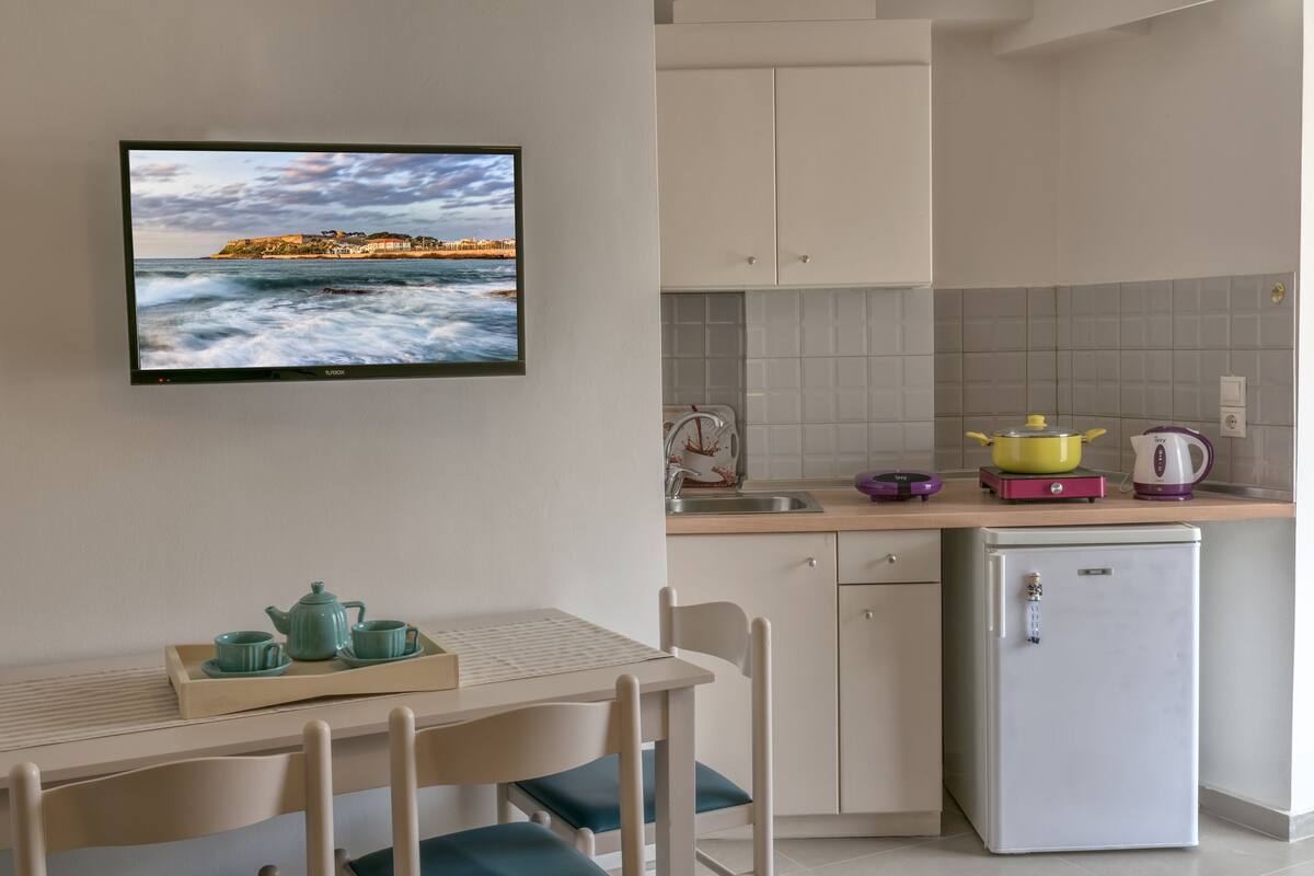 A small kitchen with a dining area, a refrigerator, a sink, and a television displaying a scenic view.