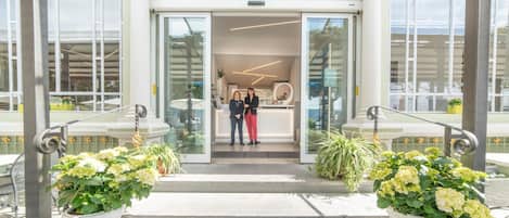Una entrada de hotel moderna con puerta de vidrio, dos plantas en macetas y una zona de recepción con escritorio y sillas.