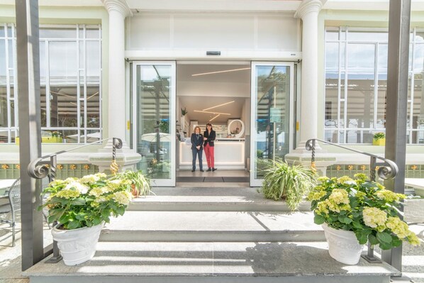 A modern hotel entrance with a glass door, two potted plants, and a reception area with a desk and chairs. - Pesce d'Oro (Verbania)