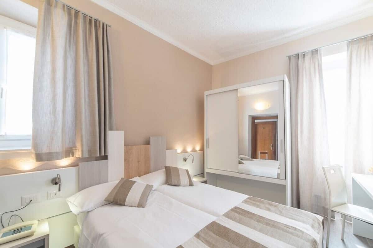 Habitación de hotel con dos camas, un escritorio, una silla, un espejo y un ventanal con cortinas.