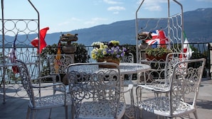 Un área de estar al aire libre con sillas de metal decorativas y una mesa, rodeada de plantas en macetas ornamentales y vistas a montañas.