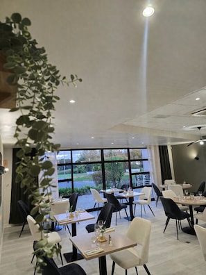 A modern restaurant with tables and chairs, large windows, and indoor plants. - Logis Hôtel Restaurant Les Fontaines (Lanvollon)