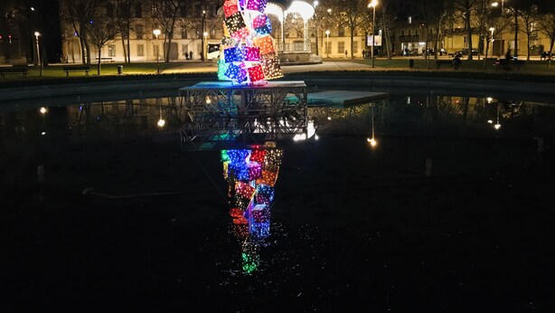 Une sculpture multicolore, illuminée de mille feux, se dresse au centre d’une fontaine, et ses couleurs se reflètent sur la surface calme de l’eau.
