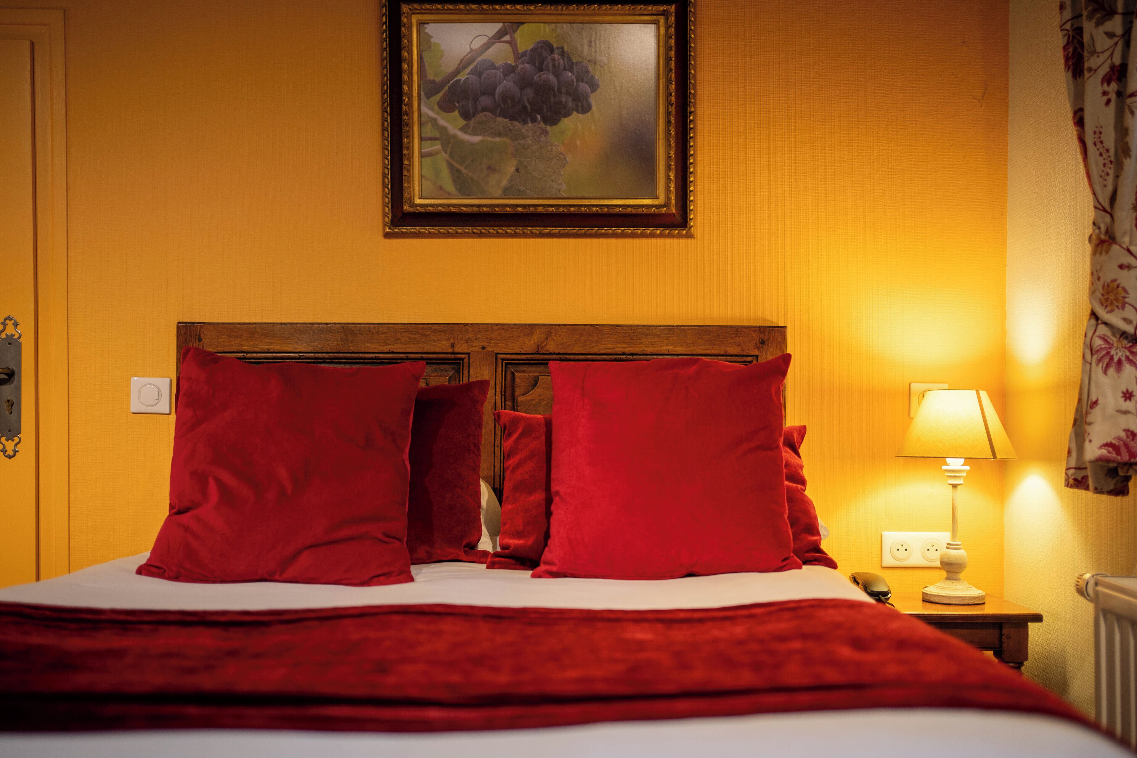 Habitación de hotel con cabecera de madera, almohadas rojas, colcha blanca, manta roja, un cuadro enmarcado en la pared, lámpara de noche y cortina con estampado floral.