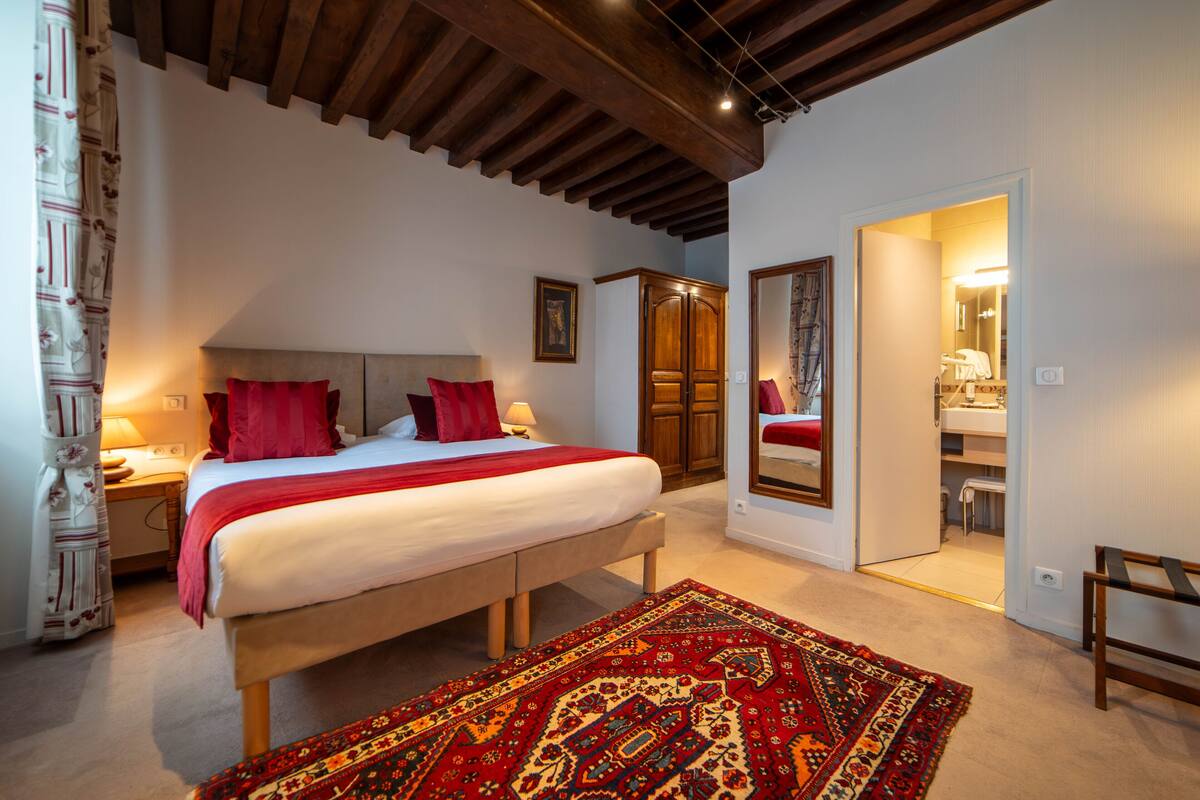 Un dormitorio con una cama grande, cabecera de madera, almohadas rojas, alfombra estampada, un armario y un espejo.