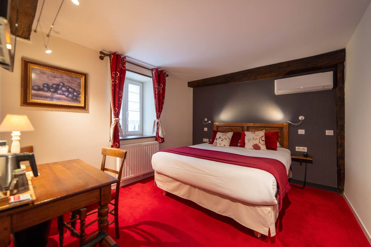 Habitación de hotel con una cama grande, un escritorio de madera, una silla, un radiador, una ventana con cortinas rojas y un cuadro en la pared.
