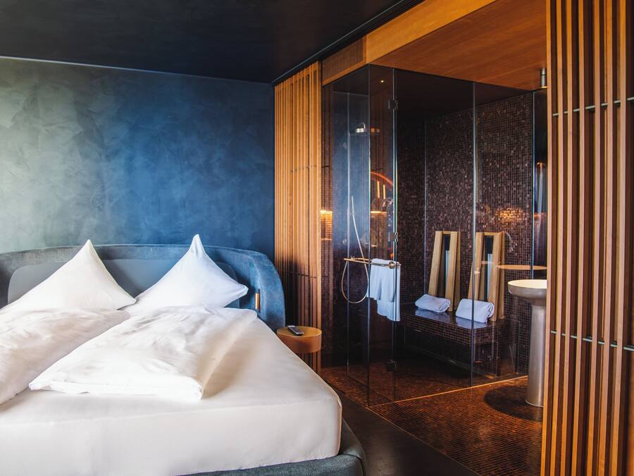 Quarto de hotel moderno com uma cama grande, área de chuveiro envidraçada e painel de parede de madeira.