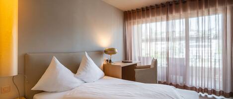 Une chambre d’hôtel avec un lit, une table de chevet, une lampe et une vue sur l’extérieur à travers des rideaux transparents.