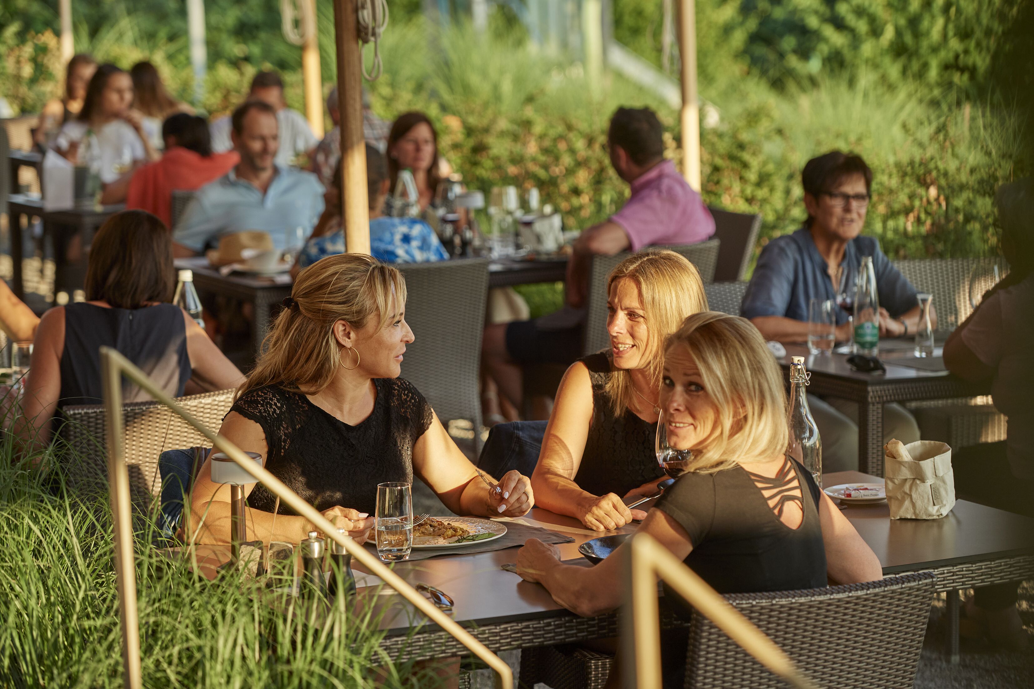 An outdoor dining area with wicker furniture and tables, surrounded by greenery.