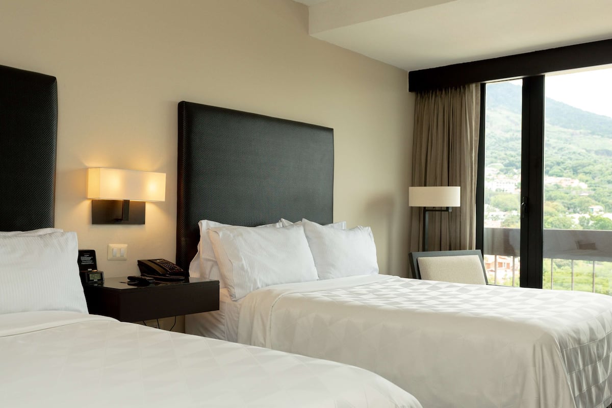 A hotel room with two beds, a nightstand with a lamp, and a view of a mountainous landscape through the window.