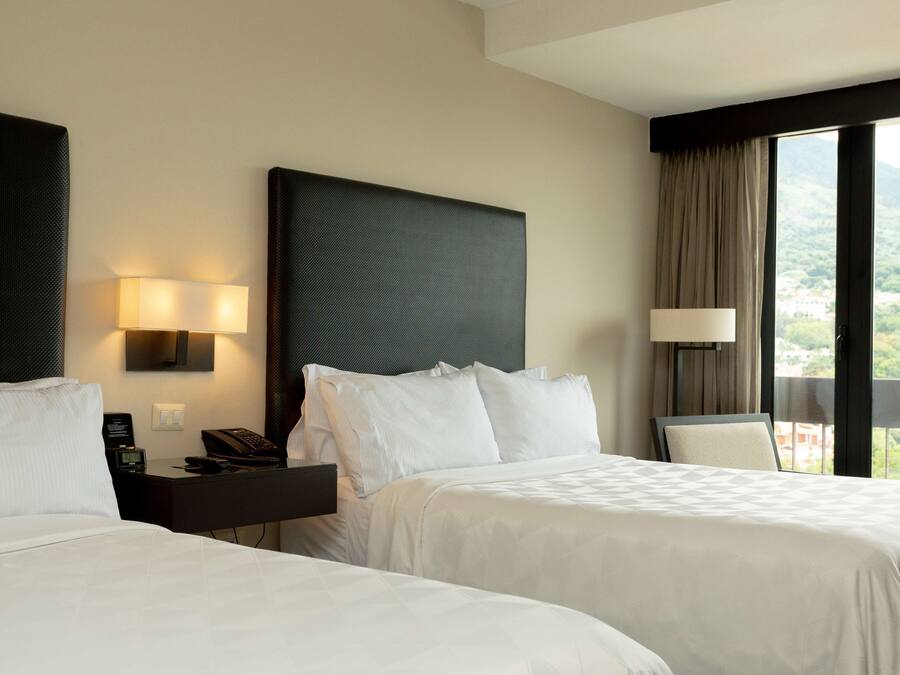 A hotel room with two beds, a nightstand with a lamp, and a view of a mountainous landscape through the window.