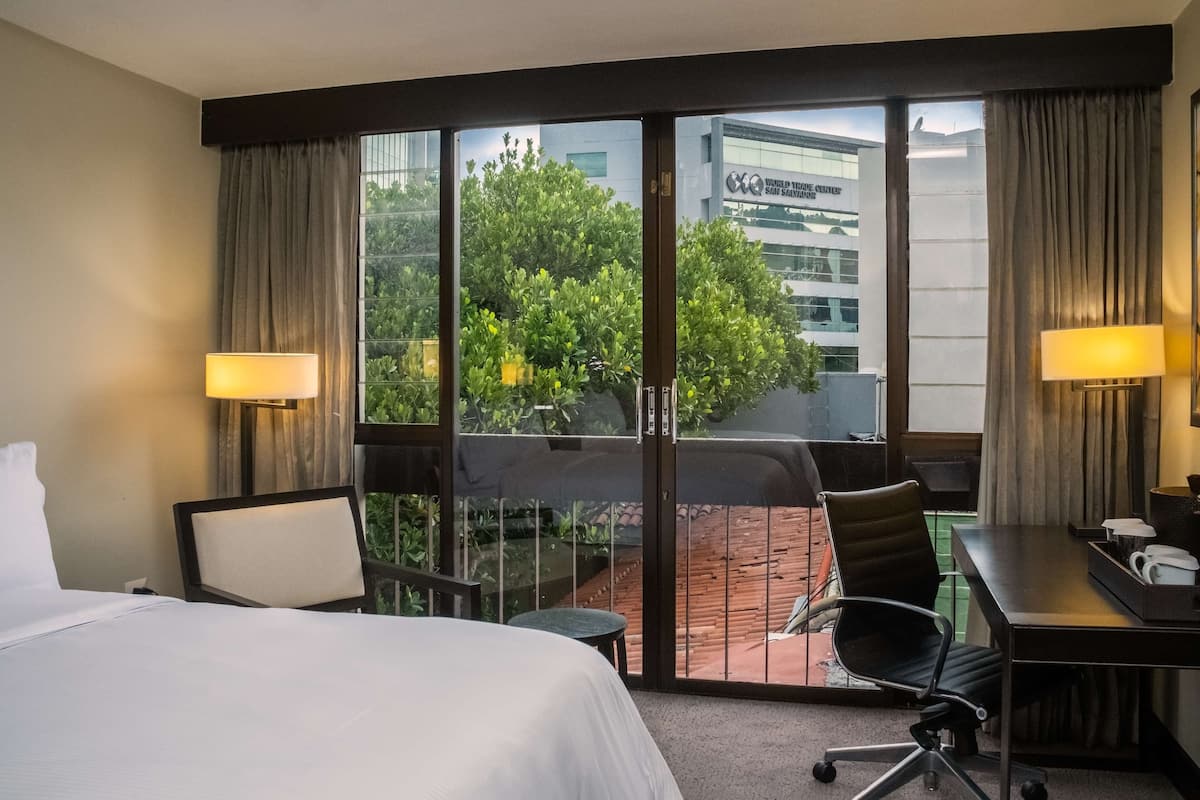 A hotel room with a bed, desk, and chair, a balcony with a view of greenery and buildings, and a large window with curtains.