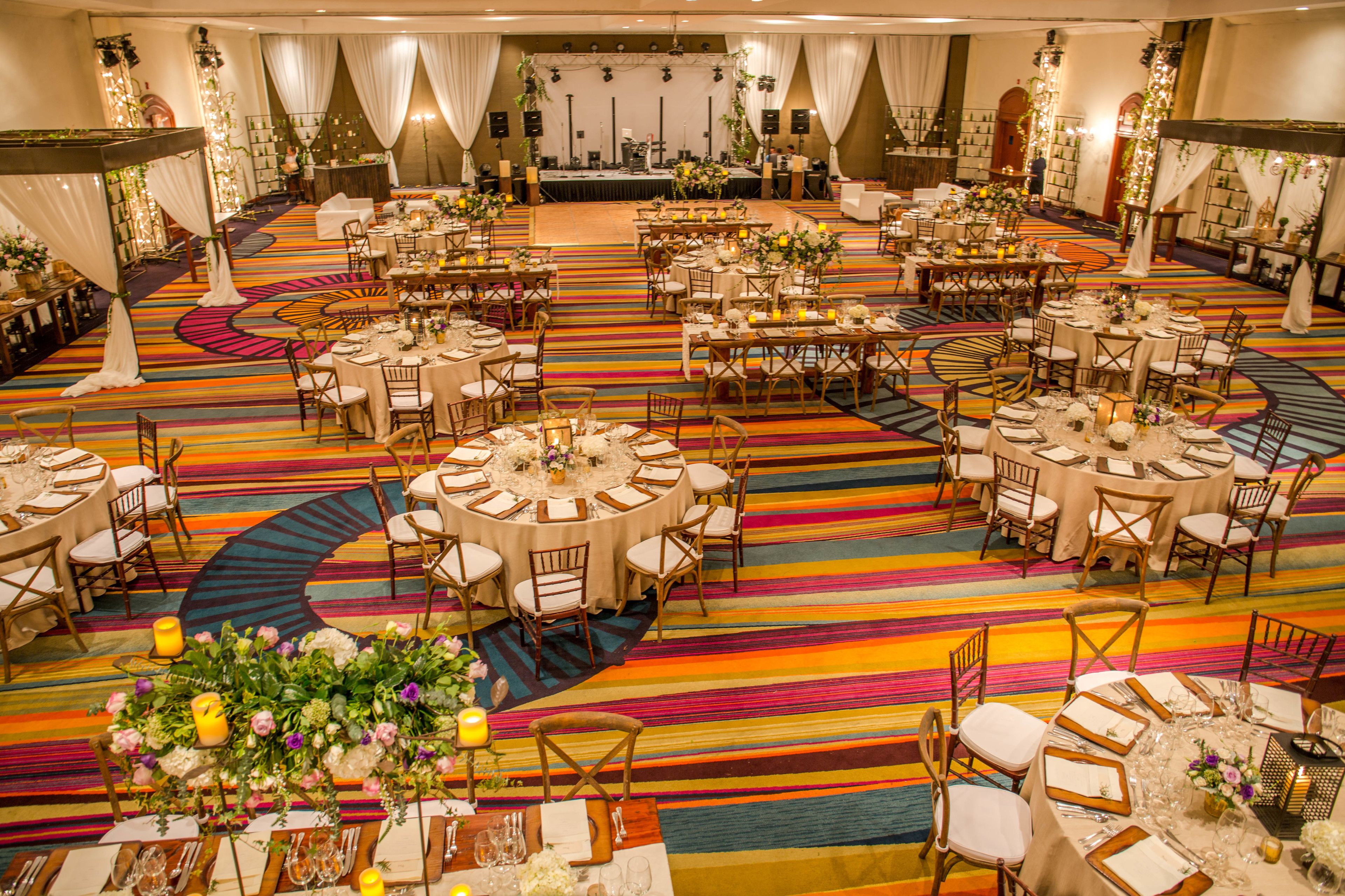 Un amplio salón de banquetes con varias mesas redondas y sillas, una alfombra a rayas de colores y centros de mesa florales.