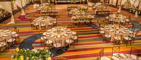 Un amplio salón de banquetes con varias mesas redondas y sillas, una alfombra a rayas de colores y centros de mesa florales.