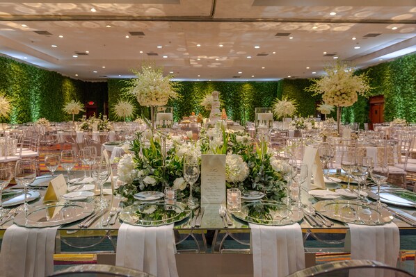 A banquet hall with round tables set for a formal event, featuring floral centerpieces and a tiered cake on the head table.