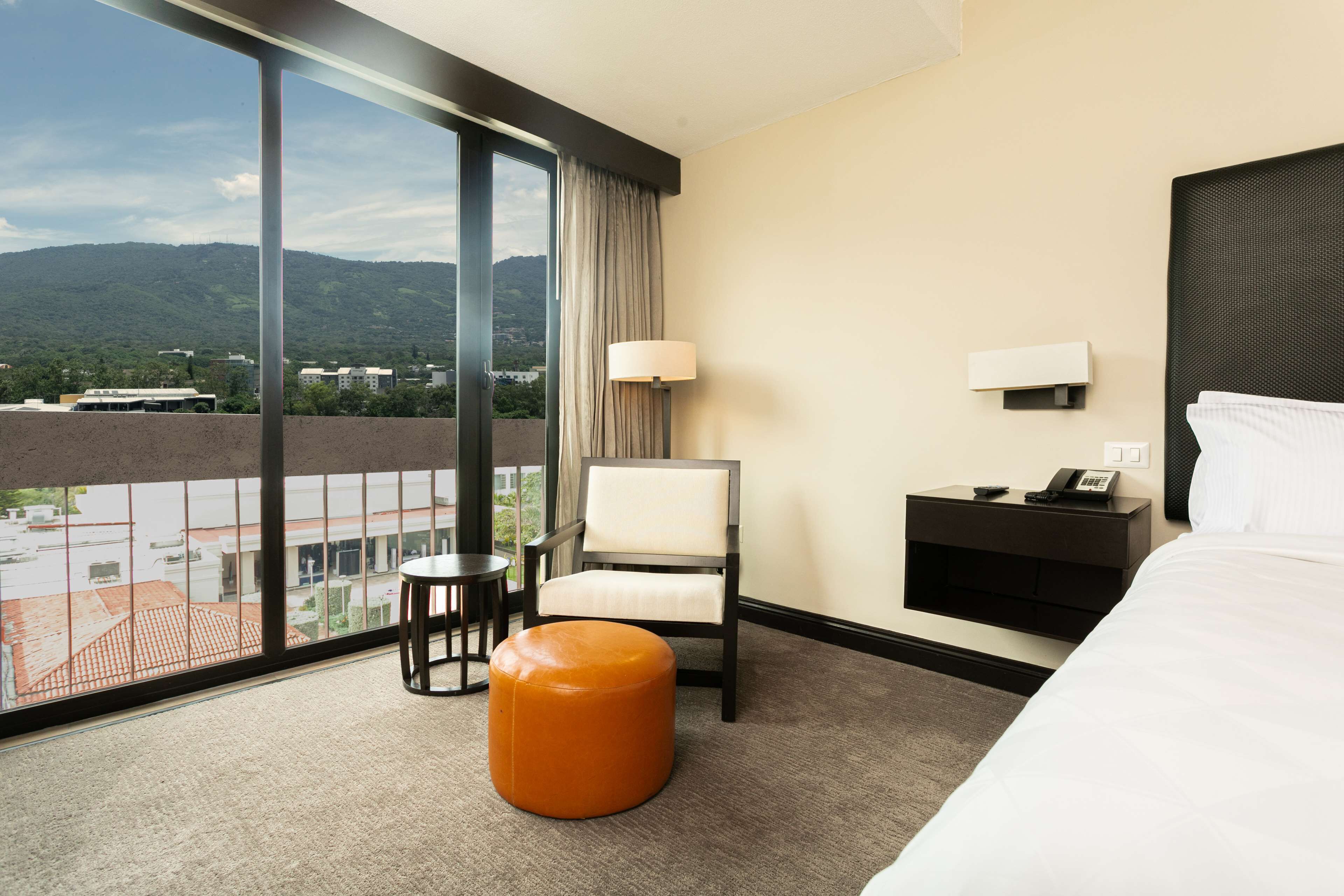 Quarto de hotel com uma janela grande, uma cama, uma cadeira, uma mesinha e uma mesa de cabeceira com abajur.
