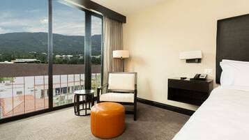 Quarto de hotel com uma janela grande, uma cama, uma cadeira, uma mesinha e uma mesa de cabeceira com abajur.