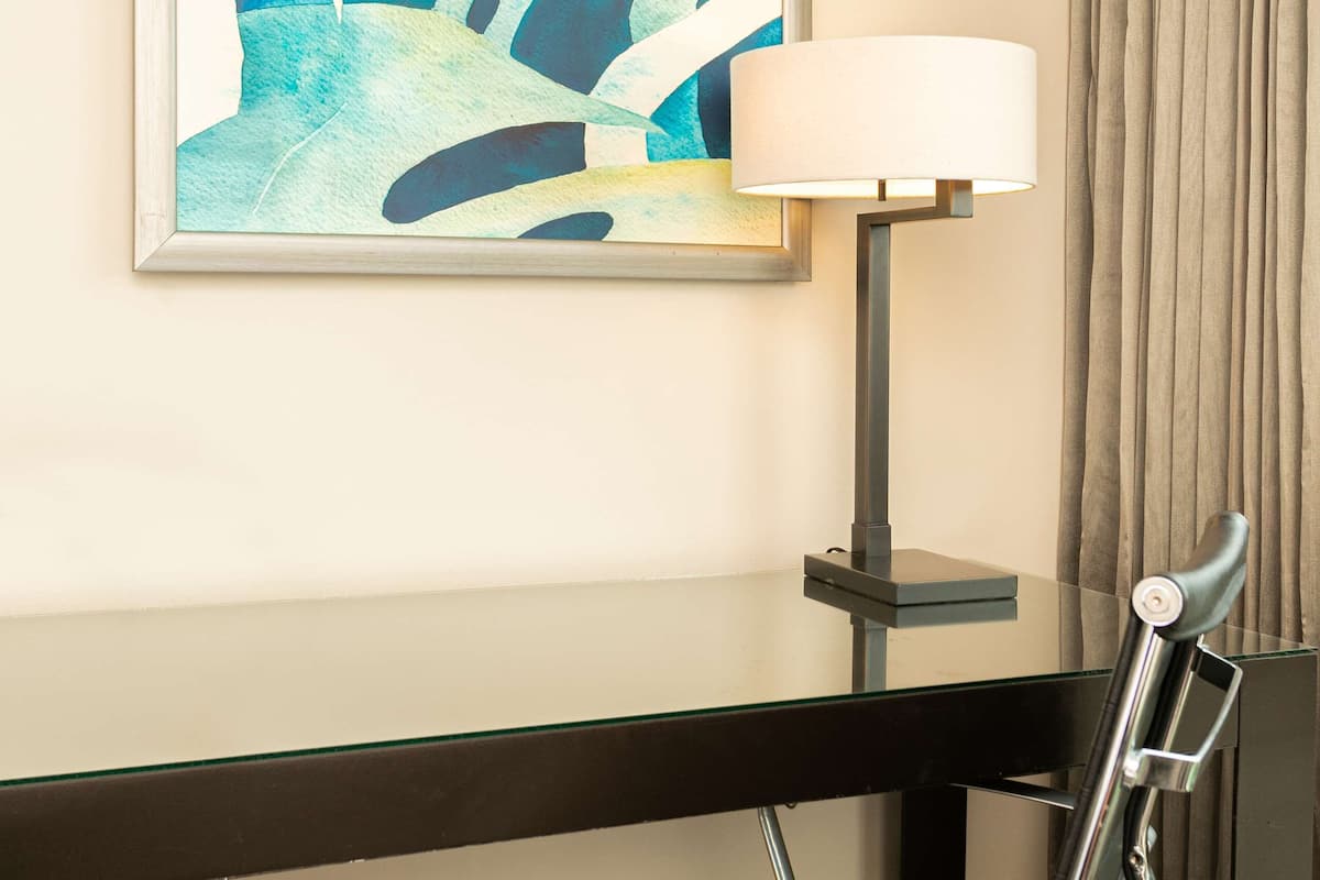 A hotel room with a desk, a chair, a lamp, and a large abstract painting on the wall.
