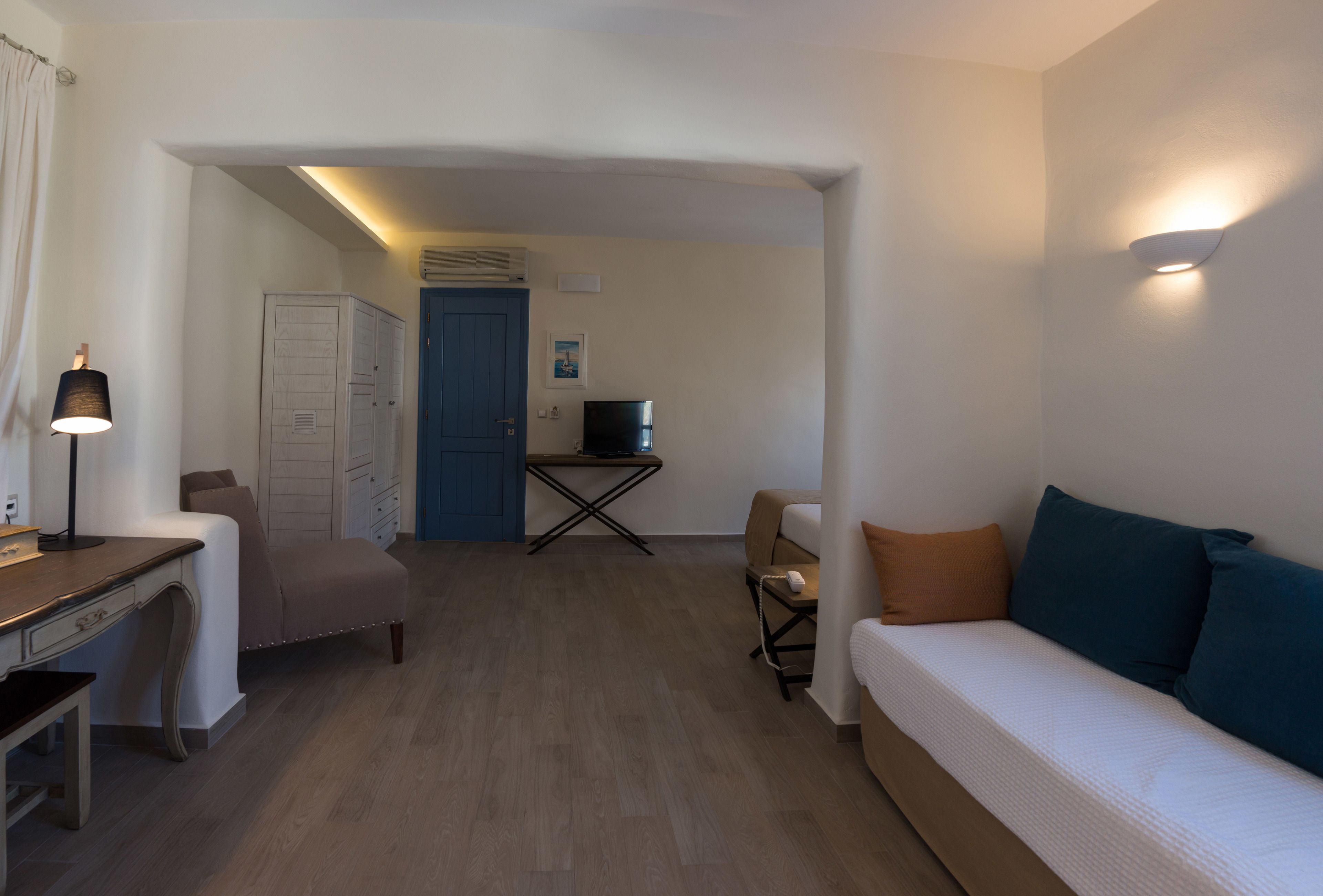 Quarto com escrivaninha, sofá, poltrona, televisão e porta.