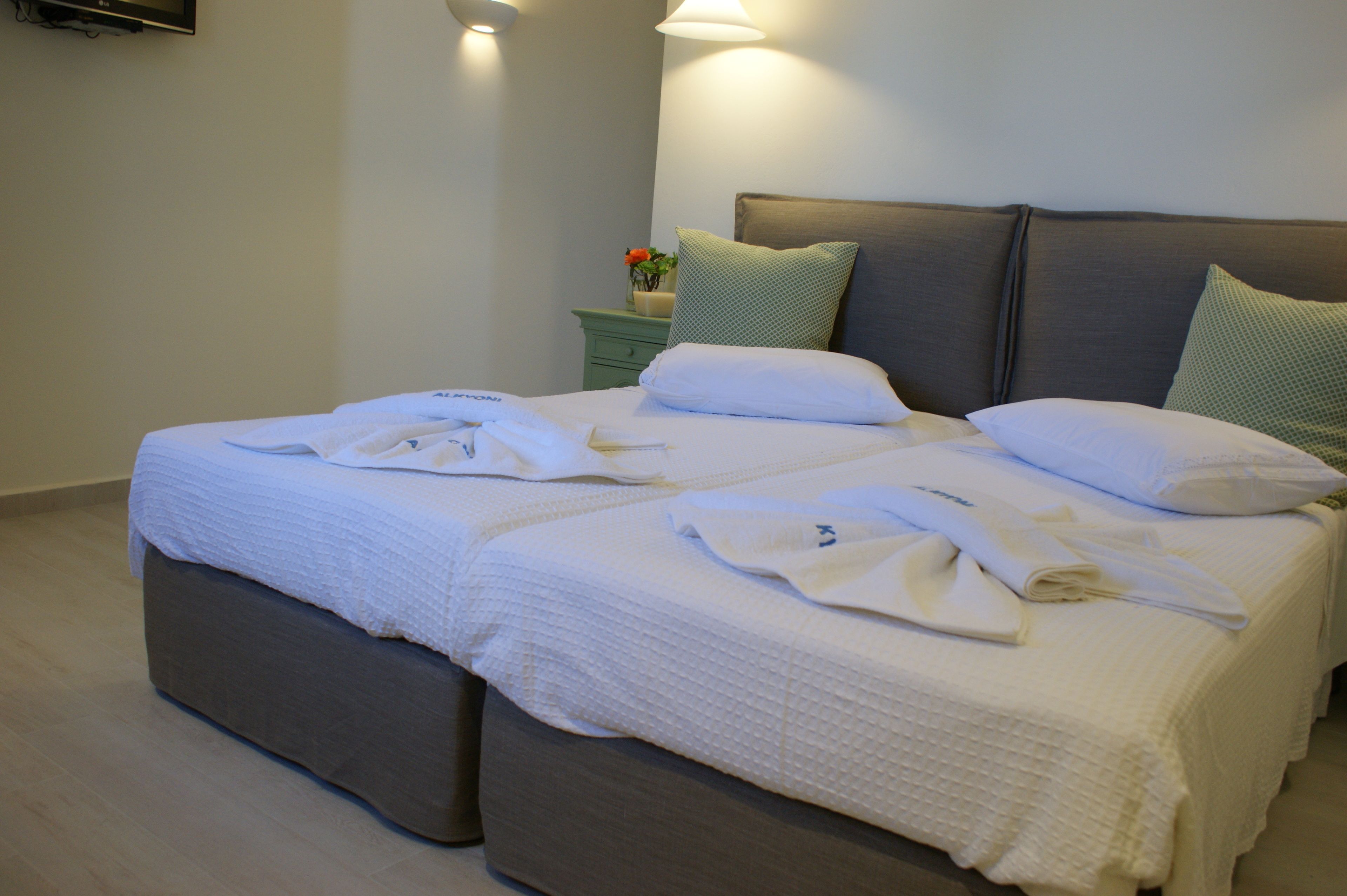 Una cama doble con sábanas y almohadas blancas, una mesita de noche con un florero y un televisor montado en la pared.