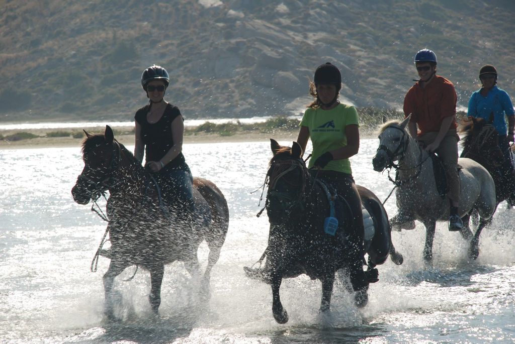 Three people riding horses in a body of water, with a mountainous background.