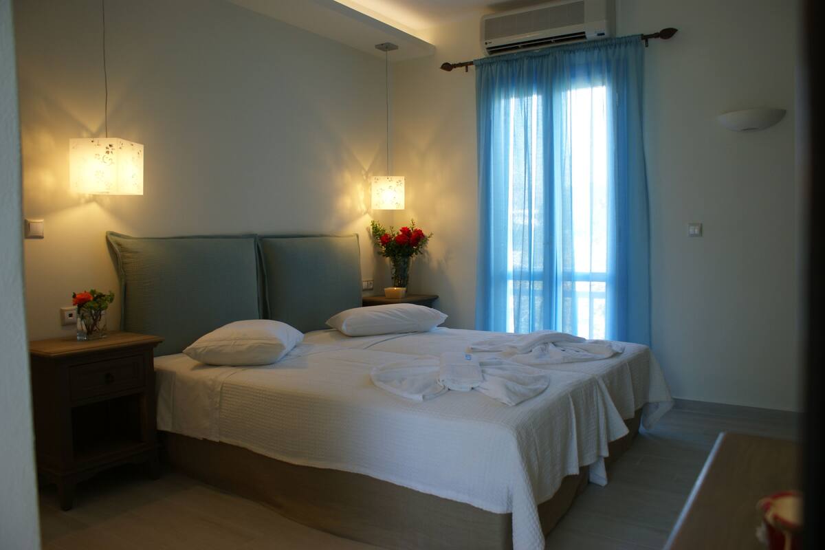 A neatly made bed with white linens, a bedside table with a vase of flowers, and a wall-mounted lamp.