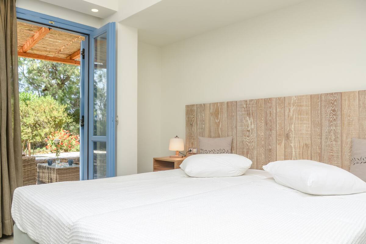 A bedroom with a large bed, a wooden headboard, a nightstand with a lamp, and a view of greenery outside.