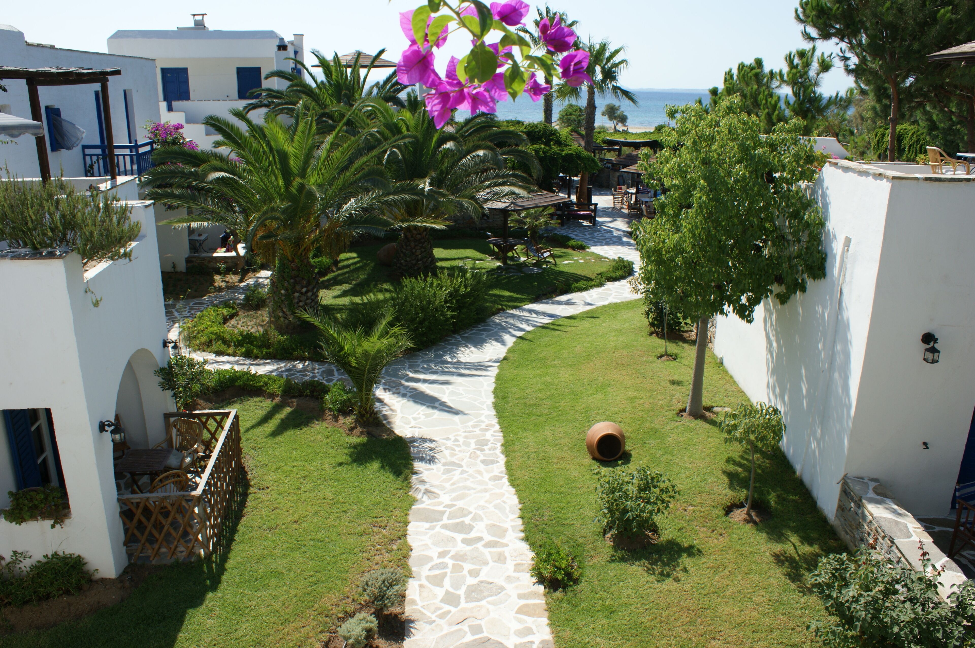A pathway leading to a pool area surrounded by white buildings with blue doors, palm trees, and a view of the sea.