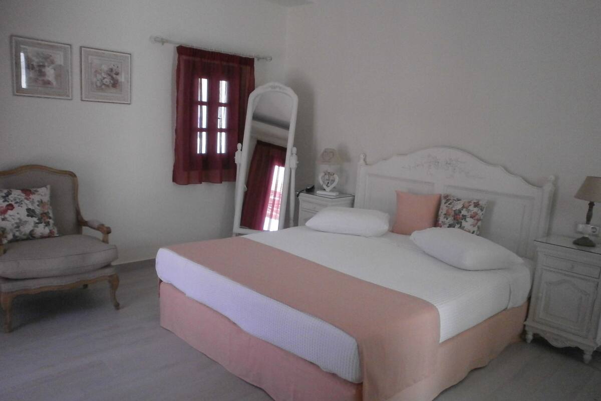A bedroom with a large bed, a chair, a nightstand, a mirror, and two framed pictures on the wall.