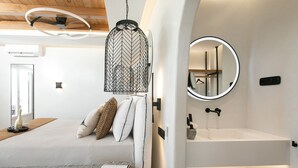 Un dormitorio moderno con cama, almohadas, una mesita de noche con una revista, una lámpara colgante y un baño con lavamanos y espejo.