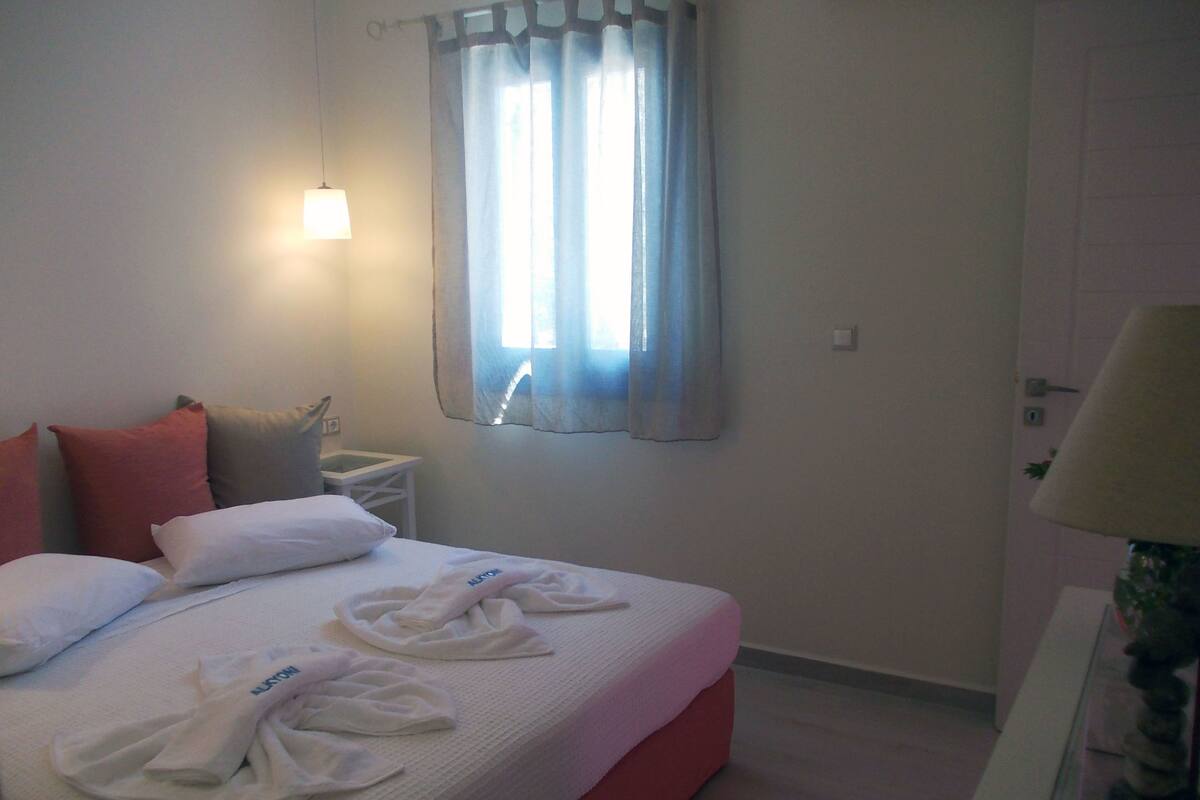 A hotel room with a bed, pillows, a nightstand, a window with curtains, and a lamp.