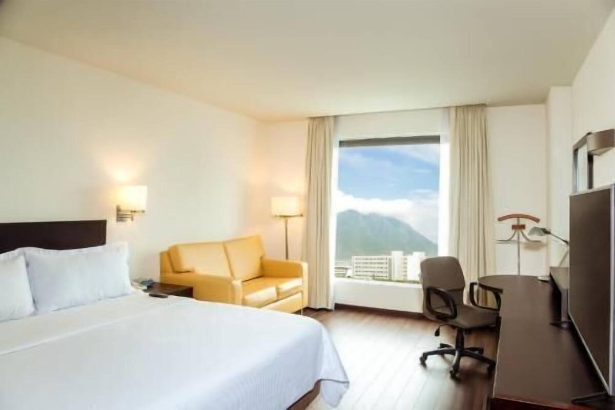 Habitación de hotel con cama, escritorio, silla, televisión y ventana con vistas a edificios y montañas.