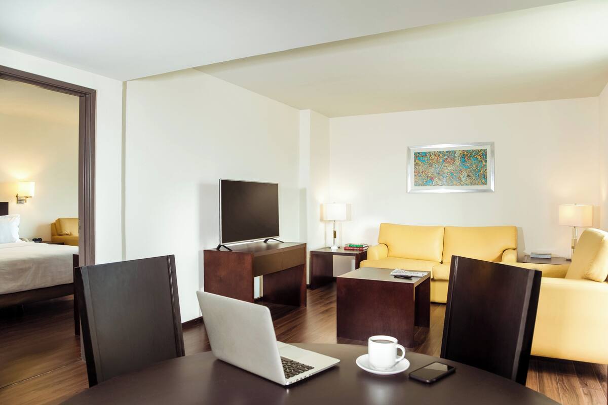 Habitación de hotel con cama, escritorio con laptop, mesa de centro, sofá y televisor.