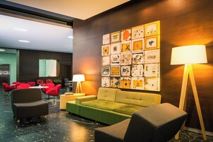 Un vestíbulo moderno con un sofá verde, sillas rojas, una lámpara de pie de madera y una pared decorada con diversas obras de arte enmarcadas.